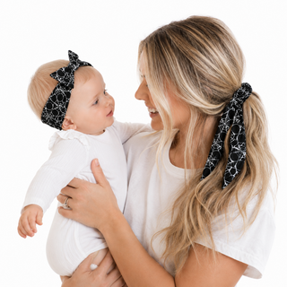 A woman holding a baby wearing matching black and white spiderweb-patterned hair bows, one in a ponytail and one as a headband, against a solid white background.
