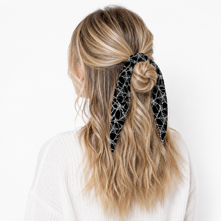 A woman shown from the back wearing a black and white spiderweb-patterned hair tie in her half-up ponytail, paired with a white knit sweater against a plain white background.